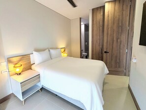 A modern bedroom with a large bed, bedside tables, a wooden wardrobe, and a bathroom visible through an open door. - Nuruma Suites by Avia Suites (Santa Marta)