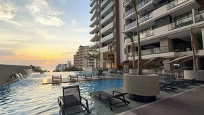 A resort pool area with lounge chairs, a water feature, and a view of a high-rise building during sunset. - Nuruma Suites by Avia Suites (Santa Marta)