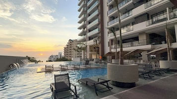 Zona de piscina en un resort con sillas de descanso, un elemento acuático y vistas a un rascacielos al atardecer.