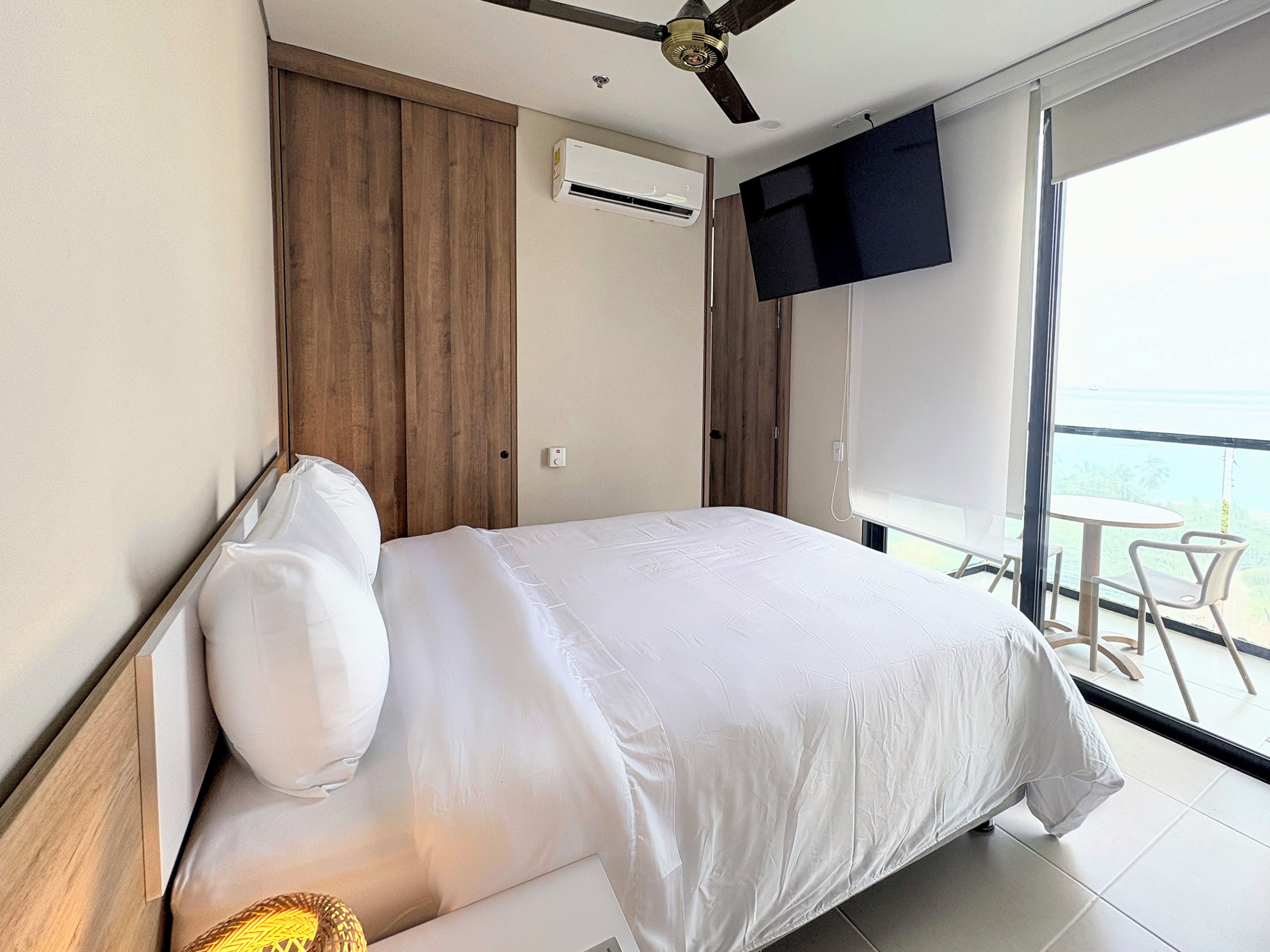 A hotel room with a bed, a ceiling fan, an air conditioning unit, a TV mounted on the wall, and a balcony with a table and chairs.