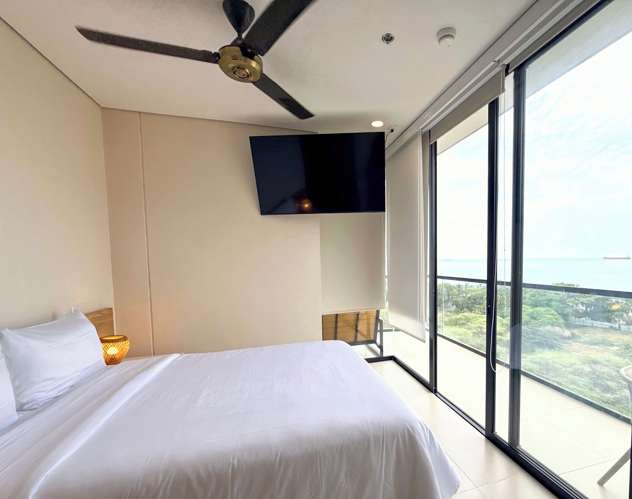 A hotel room with a bed, a ceiling fan, a TV mounted on the wall, and a large window offering a view of the sea.