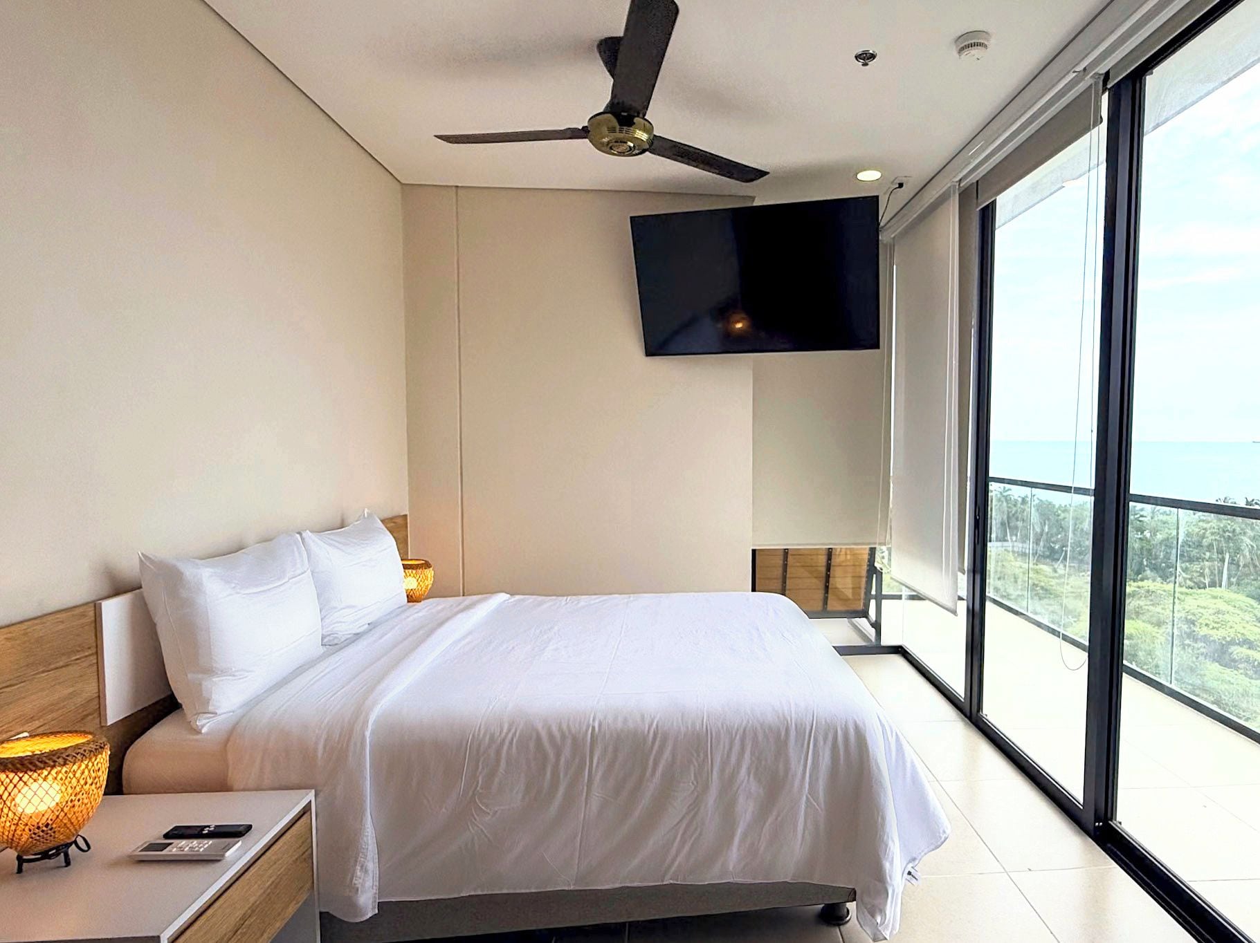 A modern hotel room with a large bed, a ceiling fan, a flat-screen TV mounted on the wall, and a balcony with a view.