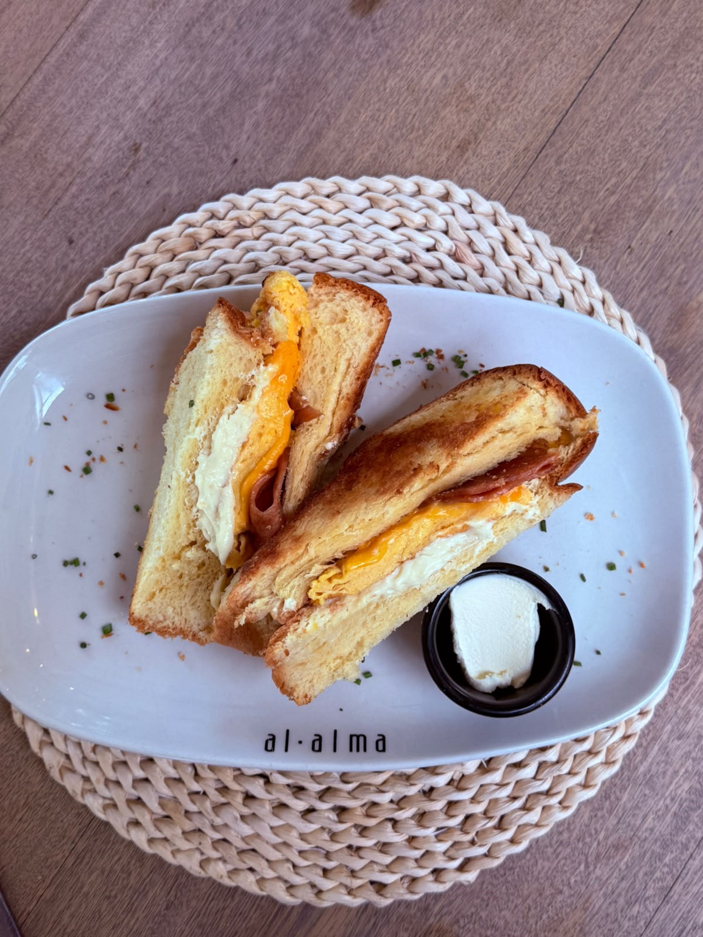 Un sándwich en un plato blanco con un recipiente pequeño de mantequilla, sobre un posavasos tejido.