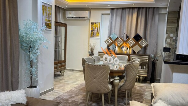 Een moderne woonkamer met een eethoek, beige banken, een kroonluchter en een groot raam.