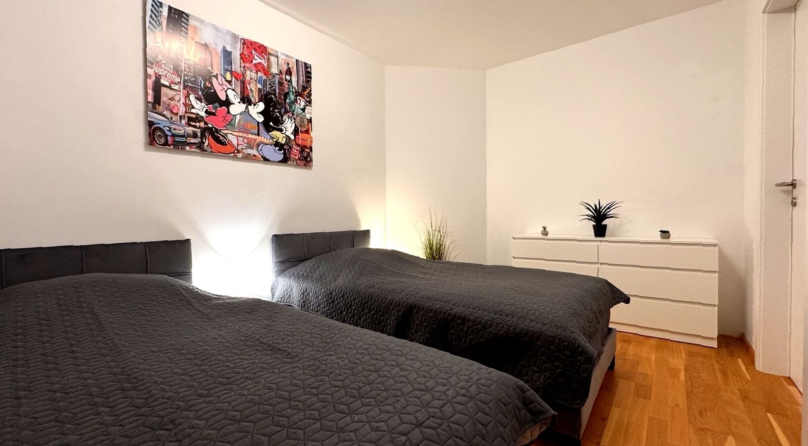 Ein Zimmer mit zwei Betten, einer weißen Kommode, einem Wandbild mit bunten Farben und einer Pflanze.