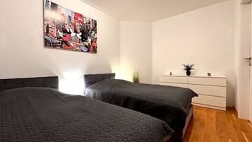 Ein Zimmer mit zwei Betten, einer weißen Kommode, einem Wandbild mit bunten Farben und einer Pflanze.