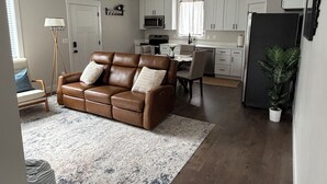 A modern living room with a brown leather sofa, a dining table, and a kitchen area with white cabinets and a black refrigerator.
