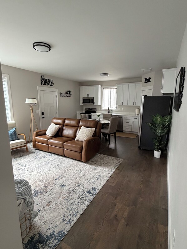 A modern living room with a brown leather sofa, a dining table, and a kitchen area with white cabinets and a black refrigerator.