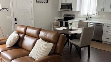 A modern living room with a brown leather sofa, a dining table, and a kitchen area with white cabinets and appliances.