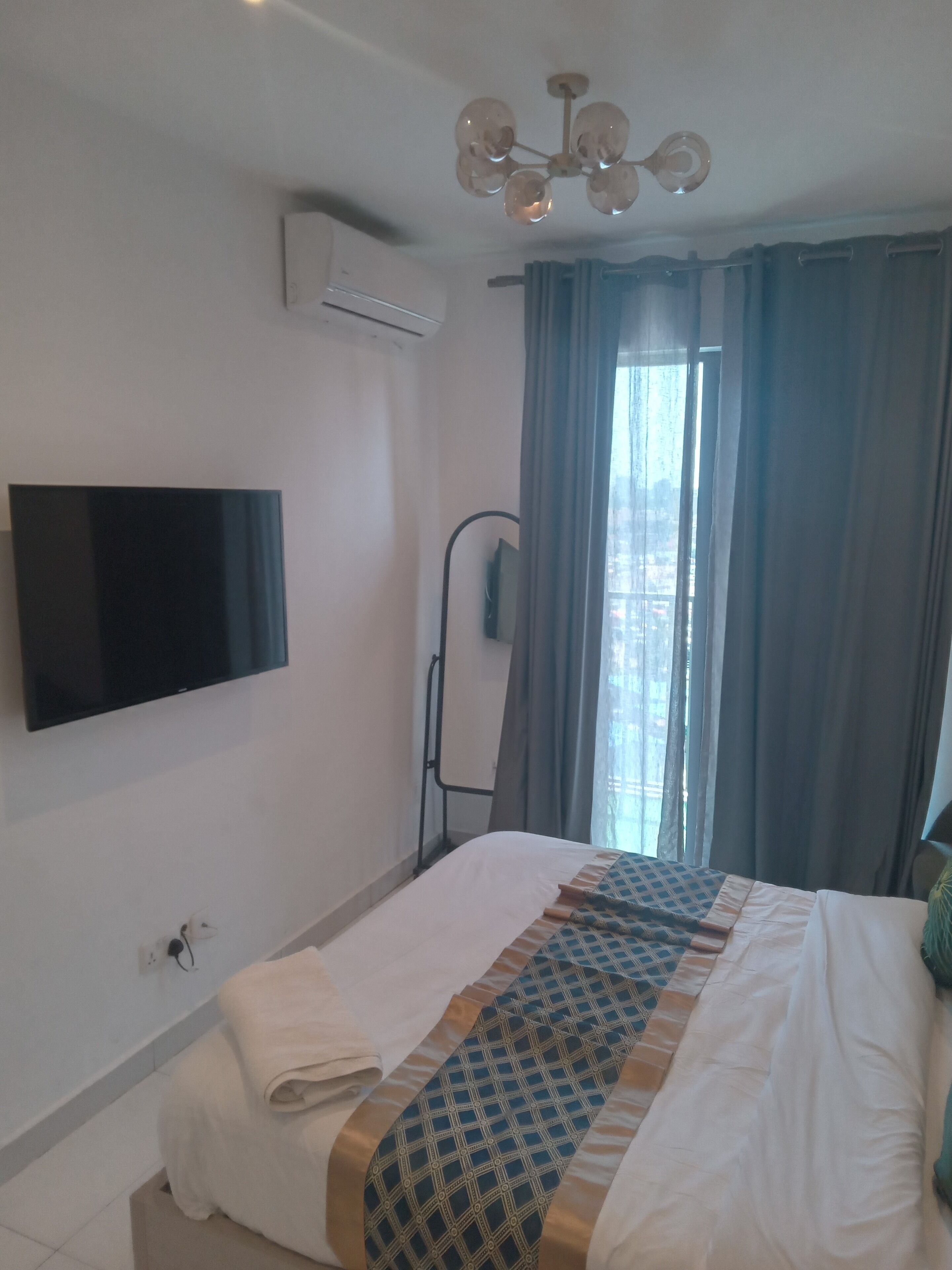 A hotel room with a bed, a flat-screen TV mounted on the wall, an air conditioning unit, a window with curtains, and a ceiling light fixture.