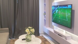 Een woonkamer met een televisie waarop een sportwedstrijd te zien is, een witte salontafel met een vaas, en een zwarte stoel.