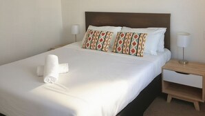 Una cama bien hecha con sábanas blancas y almohadas estampadas, una mesita de noche con lámpara, y una pared lisa con luz de techo.
