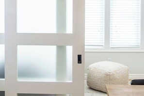Een witte deur met geëtste glaspanelen, een witte, gestructureerde poef en een houten stoel in een kamer met een raam.