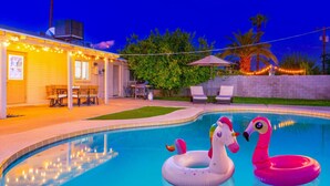 Una piscina con juguetes inflables, un patio con sillas y un edificio con luces colgantes.