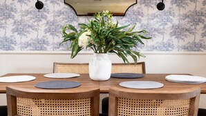 Una mesa de comedor con un jarrón blanco lleno de flores, rodeada de sillas de mimbre, en una habitación con papel tapiz estampado y un espejo en la parte superior.