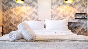 Una cama bien hecha con sábanas blancas y una toalla enrollada, dos lámparas colgantes negras y una pared con estampado floral.