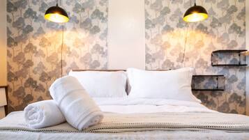 Una cama bien hecha con sábanas blancas y una toalla enrollada, dos lámparas colgantes negras y una pared con estampado floral.