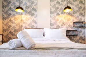 Ein ordentlich bezogenes Bett mit weißen Leinen und einem aufgerollten Handtuch, zwei schwarze Pendelleuchten und eine Wand mit Blumenmuster.