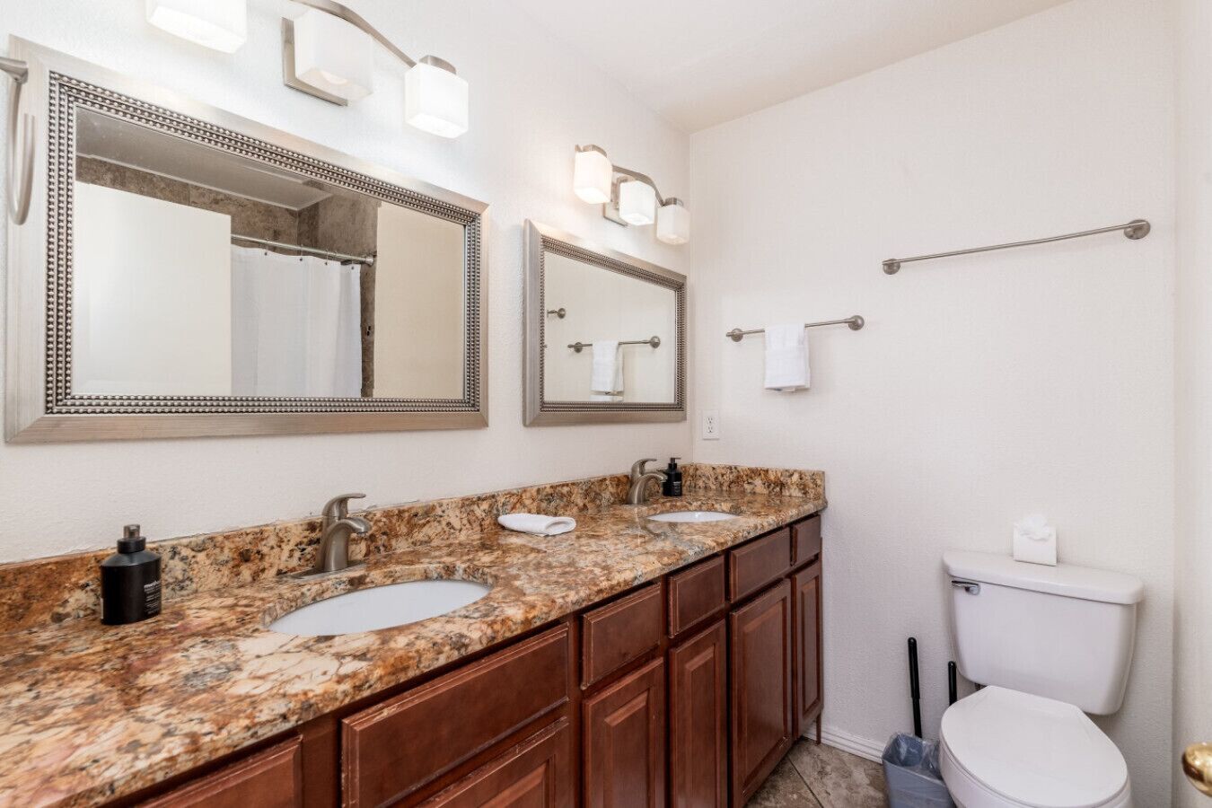 Une salle de bain dotée d’un comptoir en granit, de deux lavabos, de toilettes et de deux miroirs fixés au mur, chacun muni d’un luminaire.