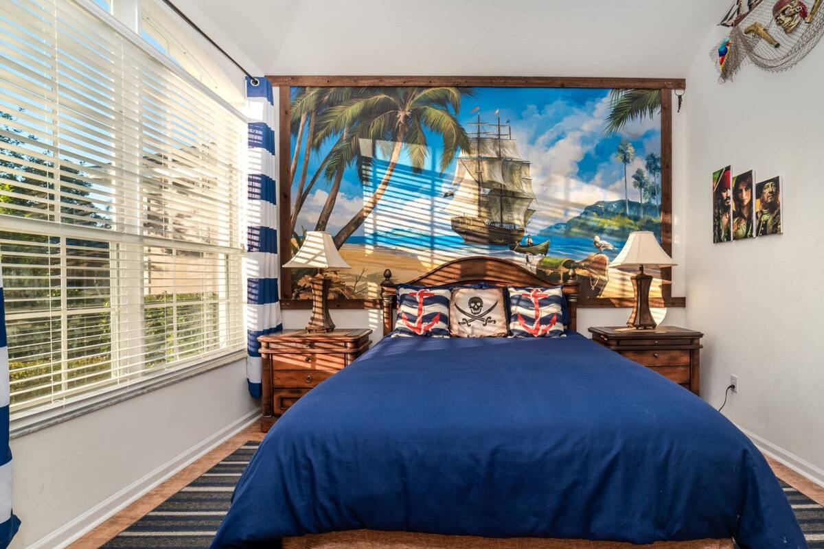 Une chambre avec un grand lit, une fresque représentant un navire et des palmiers, une fenêtre avec des stores et une porte ornée d’un miroir rond.
