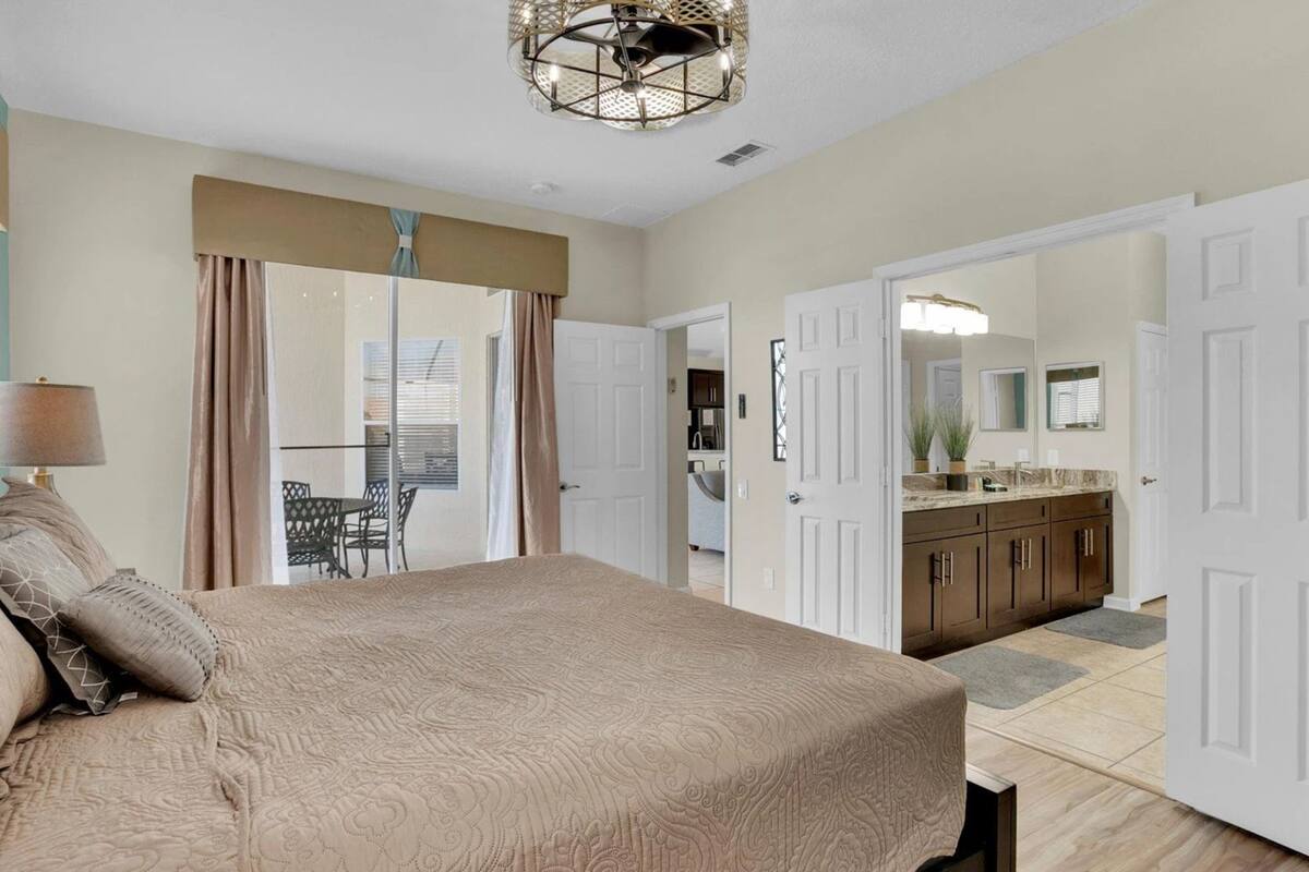 Une chambre avec un grand lit, un lustre, une porte donnant sur un balcon et un coin cuisine.