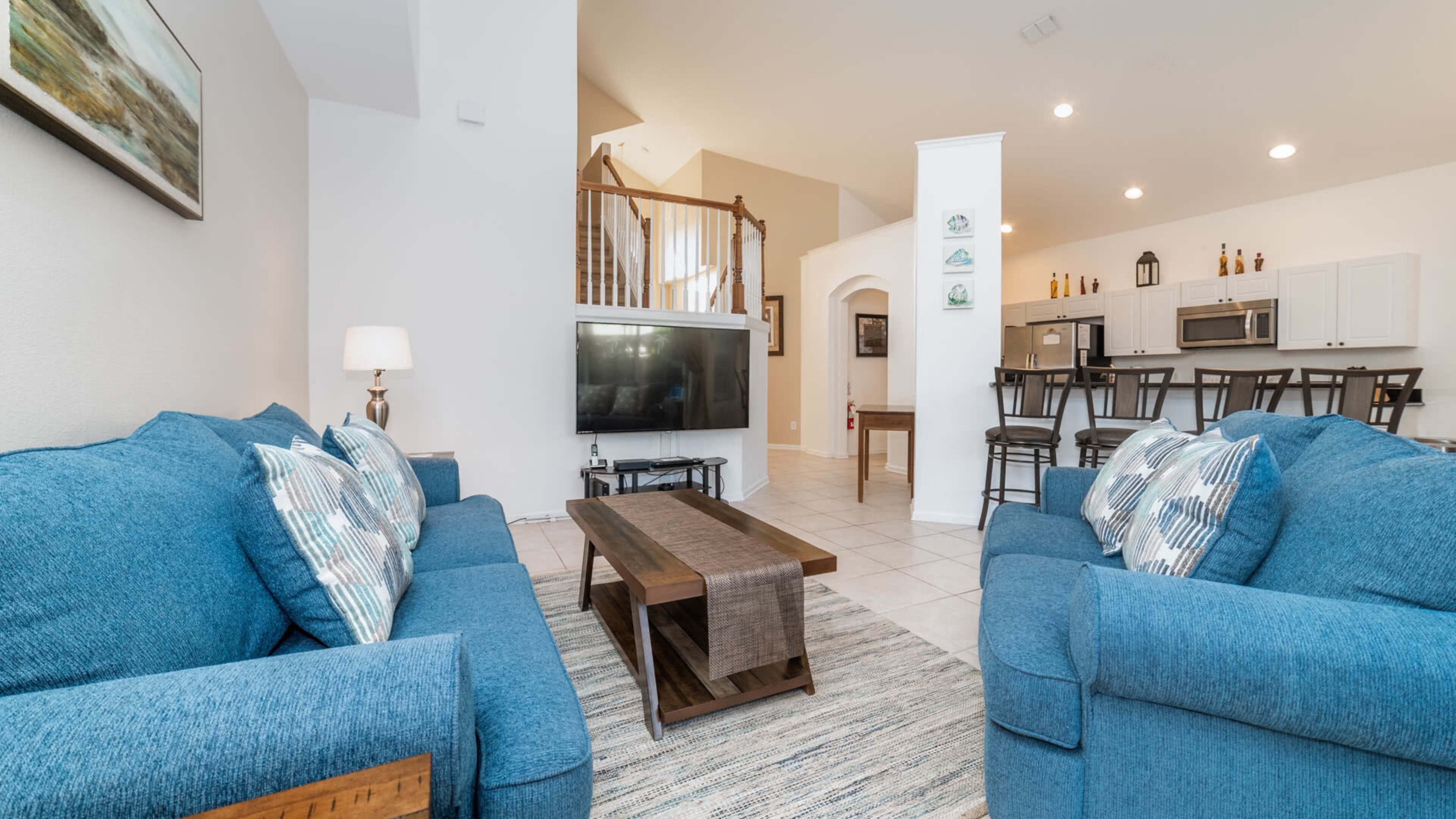 A living room with two blue sofas, a coffee table, a TV, and a staircase leading to a kitchen area.