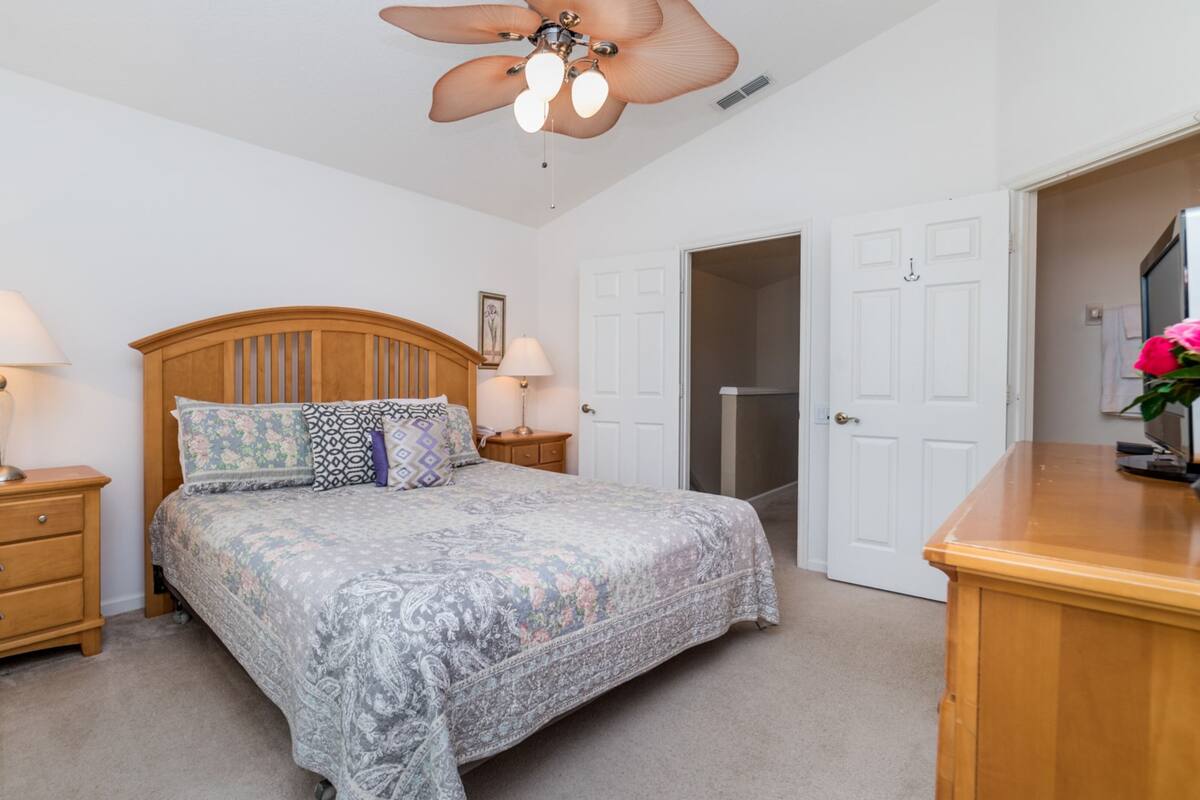 Une chambre à coucher comprenant un lit, deux tables de chevet avec des lampes, une commode en bois, un ventilateur de plafond et une porte donnant sur une autre pièce.