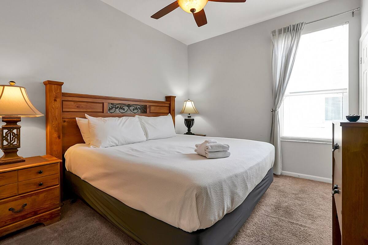 Un lit bien fait, avec du linge de lit blanc, une tête de lit en bois, une table de chevet avec une lampe, une commode et une fenêtre avec des rideaux.