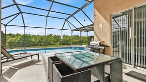 A covered outdoor swimming pool with a glass table and chairs, a grill, and a view of the forest.