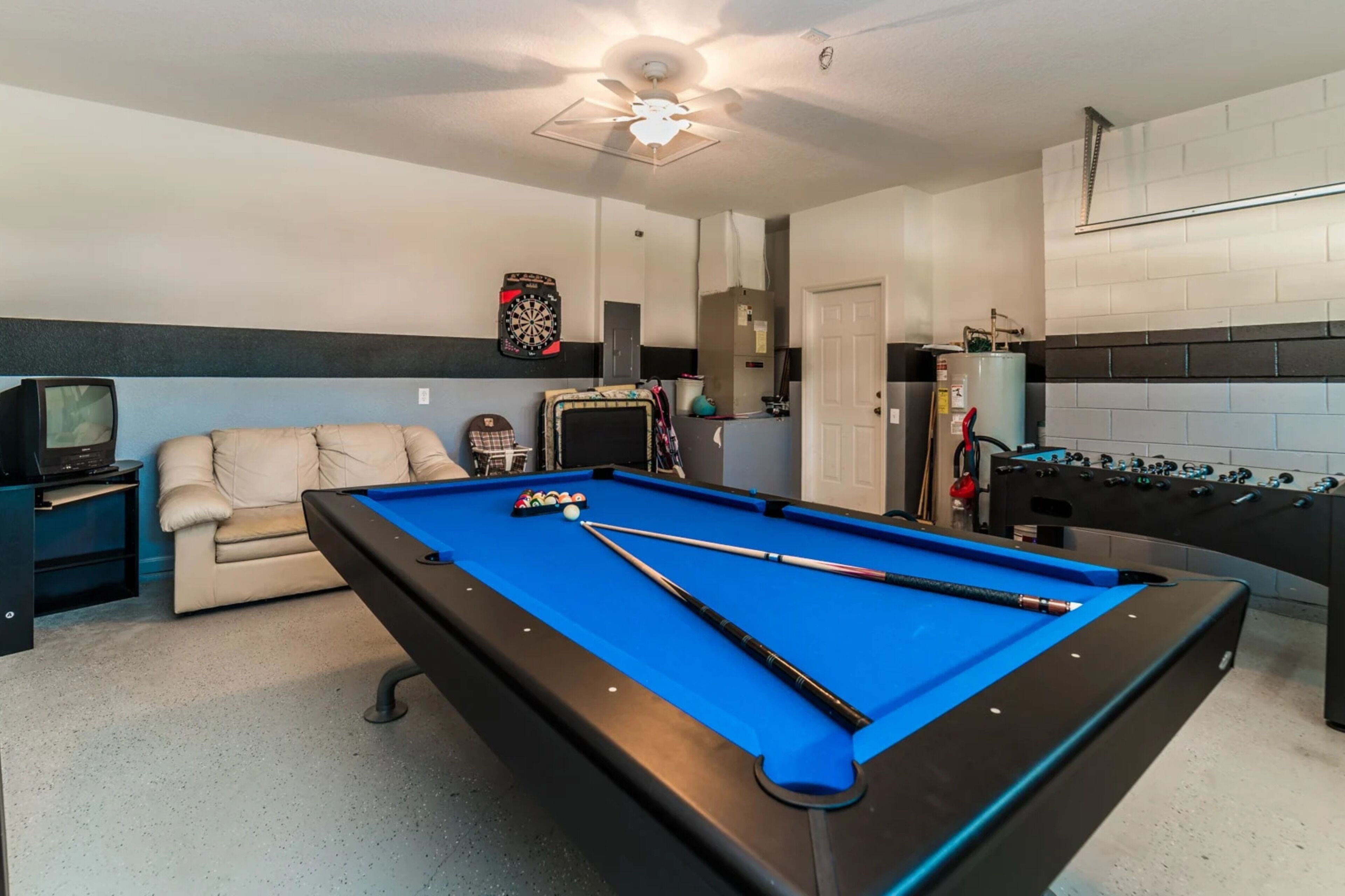 Une table de billard avec des queues de billard, un canapé, un jeu de fléchettes et une table de baby-foot dans une pièce dotée d’un ventilateur de plafond et d’un téléviseur.