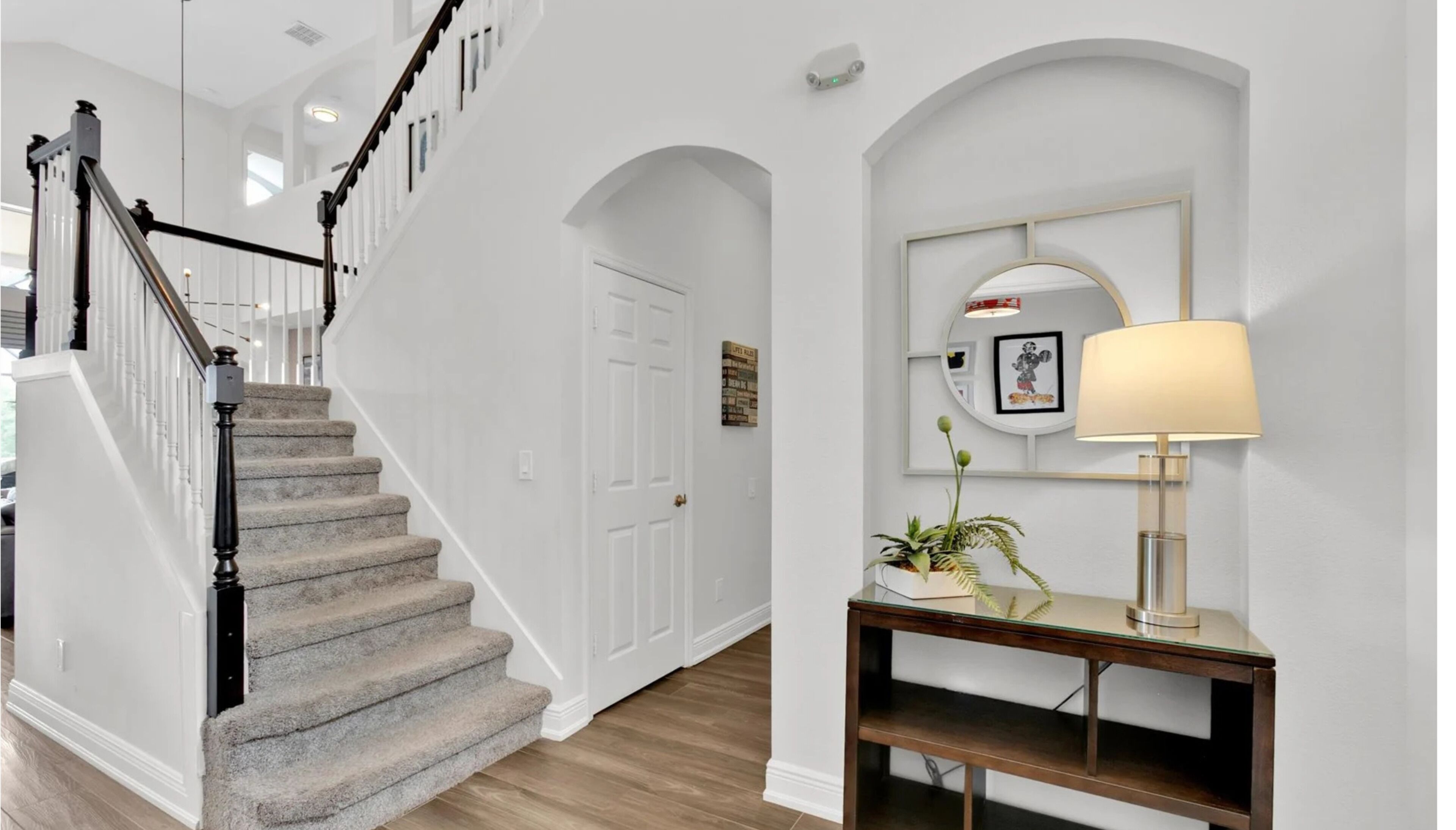 A staircase with a handrail, a white door, a console table with a lamp, and a wall-mounted mirror.