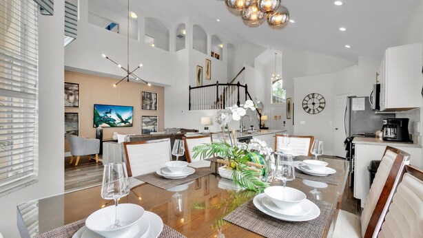 A dining area with a large table set for six, surrounded by chairs, a chandelier, and a view of the living room.