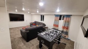 A living room with a sofa, a foosball table, a flat-screen TV, and a window with curtains.