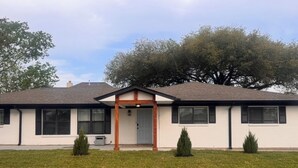 A single-story house with a front porch, a small lawn, and two evergreen shrubs.