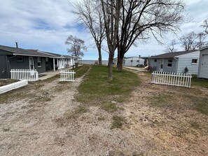 Une rangée de petits chalets à un seul étage, avec des clôtures en bois peintes en blanc, un chemin bordé de gazon et un arbre au centre.