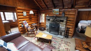 Interior de una cabaña de troncos con chimenea de piedra, una mecedora, un sofá y una alfombra estampada.