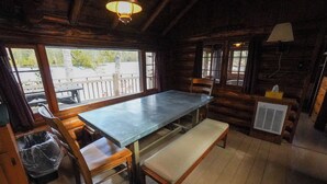 Interior de cabaña con mesa de madera, bancos, ventana con vistas, lámpara y radiador.