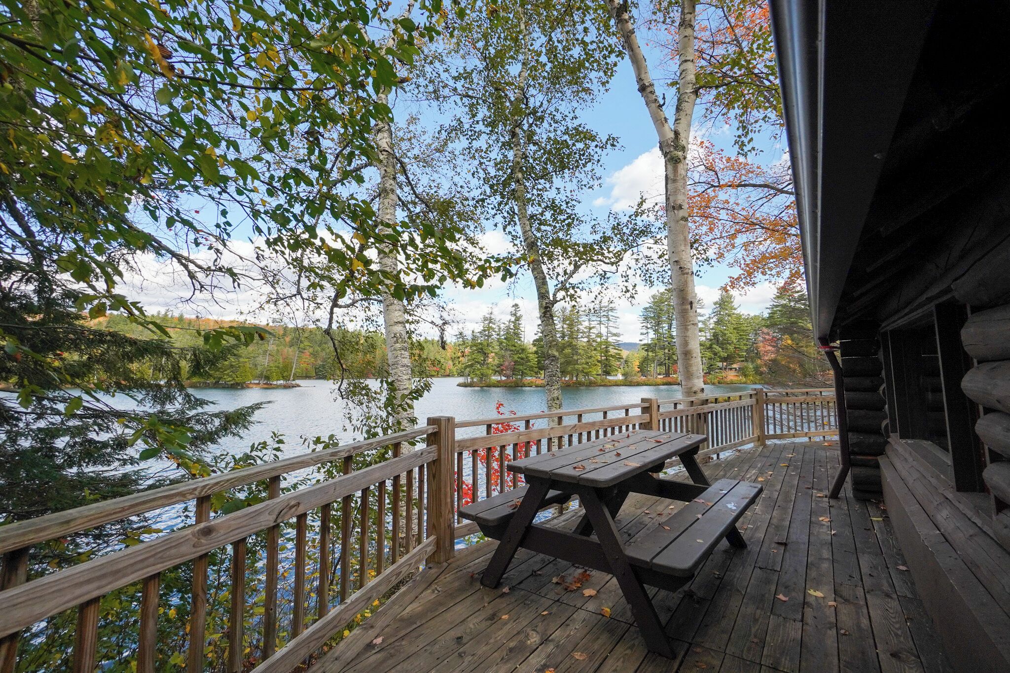 A wooden deck with picnic tables overlooking a lake, surrounded by trees and a wooden fence.