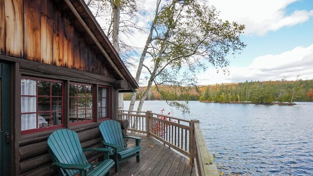 Una cabaña de madera con un porche, dos sillas Adirondack y vistas a un lago y bosque.