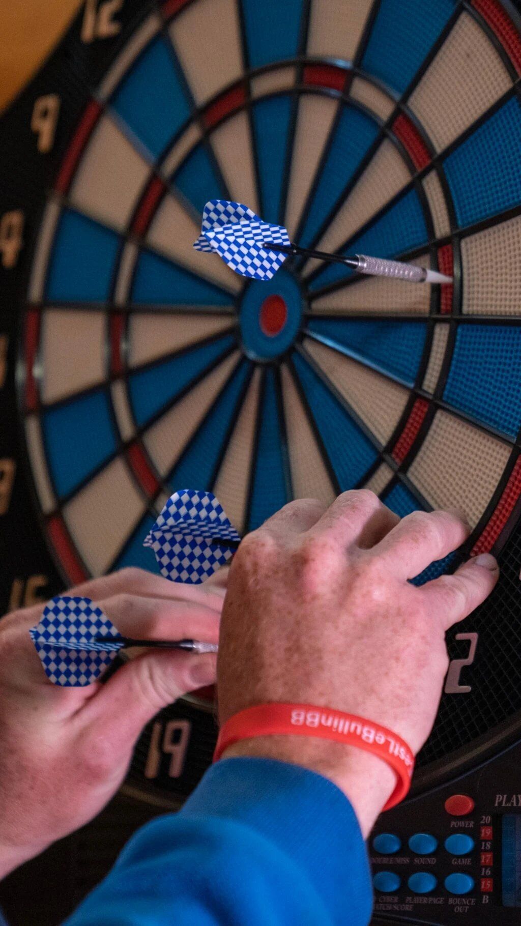 Un tablero de dardos con tres dardos, una mano sosteniendo un dardo y una pulsera roja.