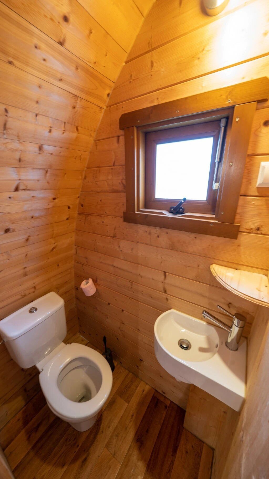 A small, wooden-paneled bathroom with a toilet, sink, and a window above the sink.