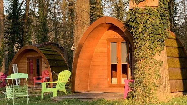 Two wooden cabins with arched entrances, surrounded by trees, and outdoor seating.