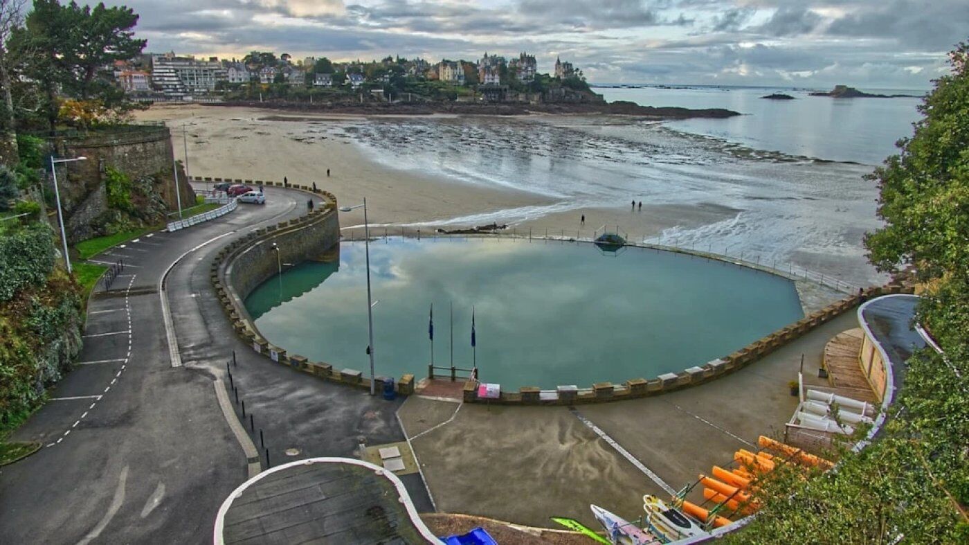 Une zone côtière avec une route sinueuse menant à une plage, un petit quai pour bateaux et un château en arrière-plan.