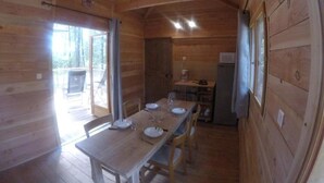 A wooden cabin interior with a dining table set for four, a kitchen area with a refrigerator, and a door leading outside.