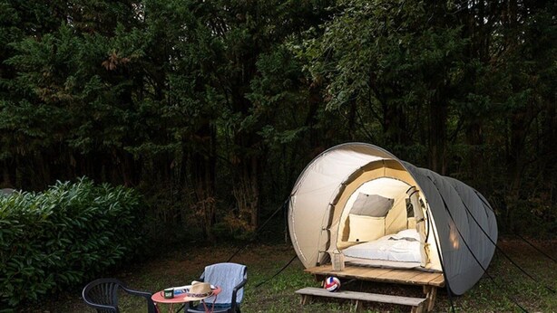 Une tente en forme de dôme, avec un lit à l’intérieur, entourée d’une forêt. On y trouve deux chaises et une petite table sur laquelle sont posés divers objets.