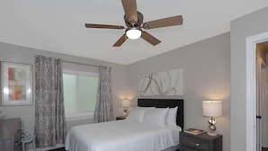 A bedroom with a bed, two bedside tables with lamps, a ceiling fan, a window with curtains, and a wall art piece. - Premium 2BR Retreat by St. Francis (Columbus)