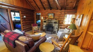 Ein Holzblockhaus-Interieur mit Kamin aus Stein, einer Ledersofa, Holzstühlen und einem Bett im Hintergrund.