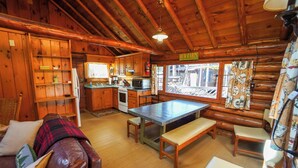 Ein Blockhaus-Interieur mit einem Holzespeisetisch, Bänken, einer Couch, einer Küchenzeile und Blick durch das Fenster.