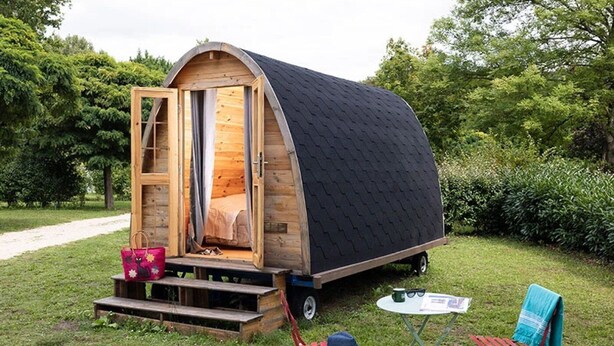 Una casa móvil pequeña de madera con techo curvo, que tiene una cama en su interior, una mesa pequeña y dos sillas afuera.