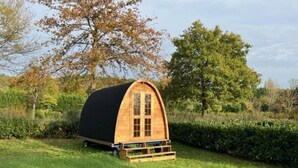 Una pequeña estructura de madera con techo negro, de forma arqueada, ubicada en un campo cubierto de césped con árboles al fondo.